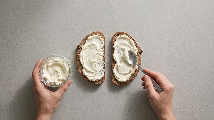 Zwei Hände verstreichen mit einem Löffel Frischkäse dick auf zwei Brotscheiben auf einer hellgrauen Arbeitsplatte.