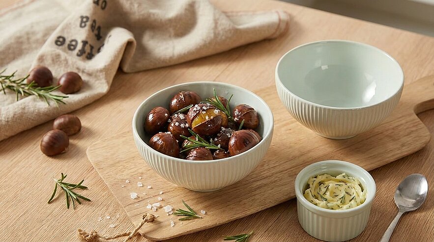 Schale mit glasierten Maronen, garniert mit Rosmarin und grobem Meersalz, auf einem Holzbrett neben einer kleinen Schale mit Kräuterbutter und einer leeren Schüssel.
