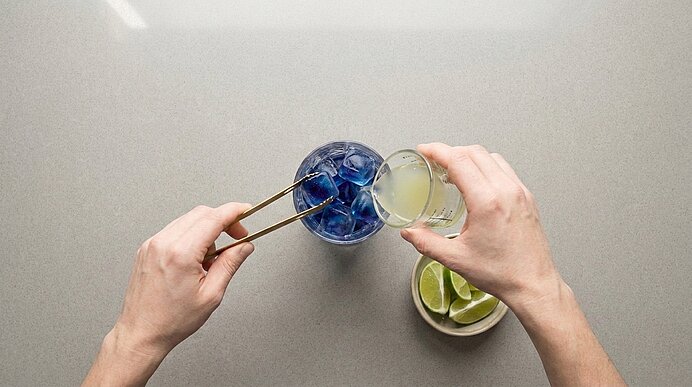 Zwei Hände bereiten ein Getränk: mit einer Zange werden blaue Eiswürfel in ein Glas gelegt, während eine helle Flüssigkeit eingegossen wird; daneben eine Schale mit Limettenstücken.