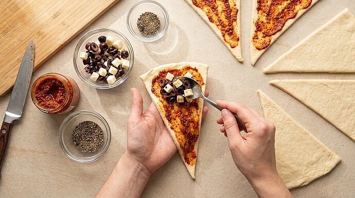 Hände füllen ein dreieckiges Pizzateigstück: auf mit Tomatensauce bestrichene Teigspitze wird eine Mischung aus schwarzen Oliven und gewürfeltem Käse gelegt; weitere Teigdreiecke und Schälchen mit Kräutern sichtbar.