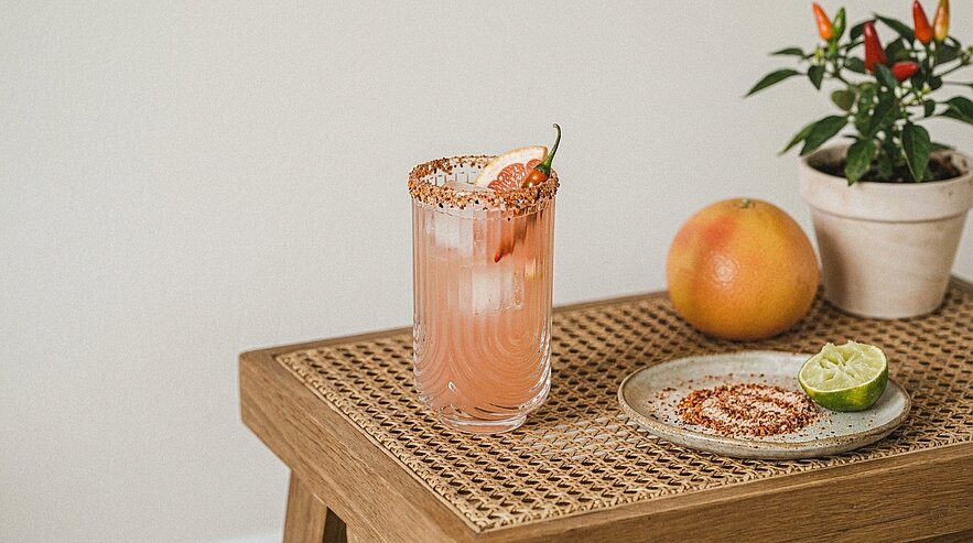 Glas mit rosa Grapefruitcocktail, Eis und gewürztem Salzrand, garniert mit Grapefruitscheibe und roter Chili; daneben Grapefruit, halbe Limette und Teller mit Gewürzsalz.