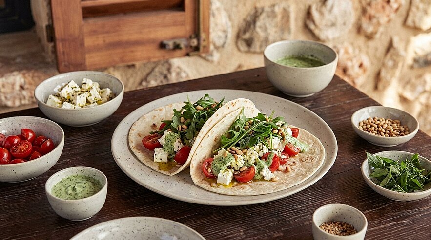 Zwei weiche Tortillas auf einem Teller, gefüllt mit Rucola, halbierten Kirschtomaten, gewürfeltem Feta und grüner Kräutersauce.