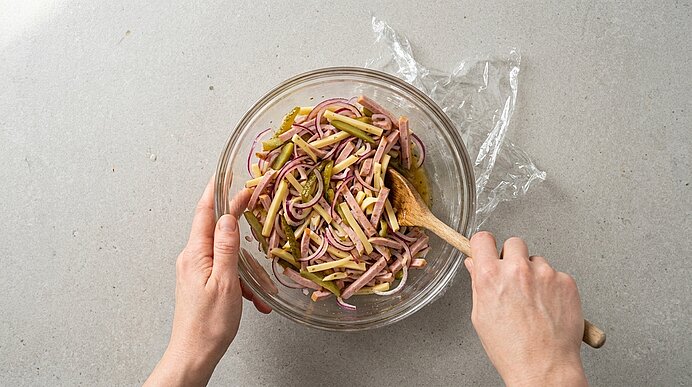 Zwei Hände rühren mit einem Holzlöffel in einer Glasschüssel einen Salat aus dünn geschnittenen Schinken- und Gurkenstreifen.
