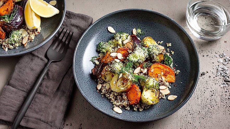 Eine dunkle Schale mit einer bunten Mischung aus geröstetem Gemüse, darunter Karotten, Brokkoli und Zwiebeln, ist auf einem Tisch angerichtet. Darunter liegt eine Schicht Quinoa, garniert mit Mandelsplittern und frischem Dill. Neben der Schale befindet sich eine Gabel und ein Wasserglas.