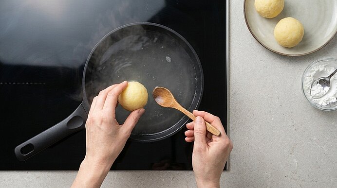 Zwei Hände setzen ein Teigbällchen mit einem Holzlöffel in eine Pfanne mit siedendem Wasser.