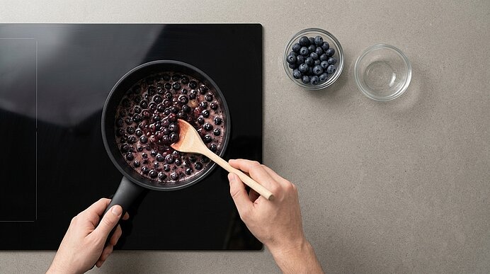 Zwei Hände rühren köchelnde Blaubeeren mit einem Holzlöffel in einem schwarzen Stieltopf auf dem Kochfeld.