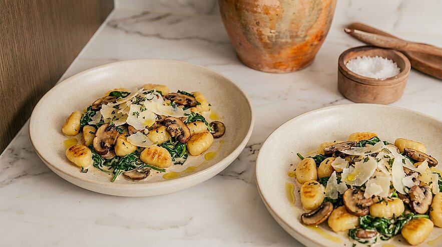 Zwei Teller Gnocchi mit gebratenen Champignons, Spinat, Olivenöl und gehobeltem Parmesan auf hellem Tisch.
