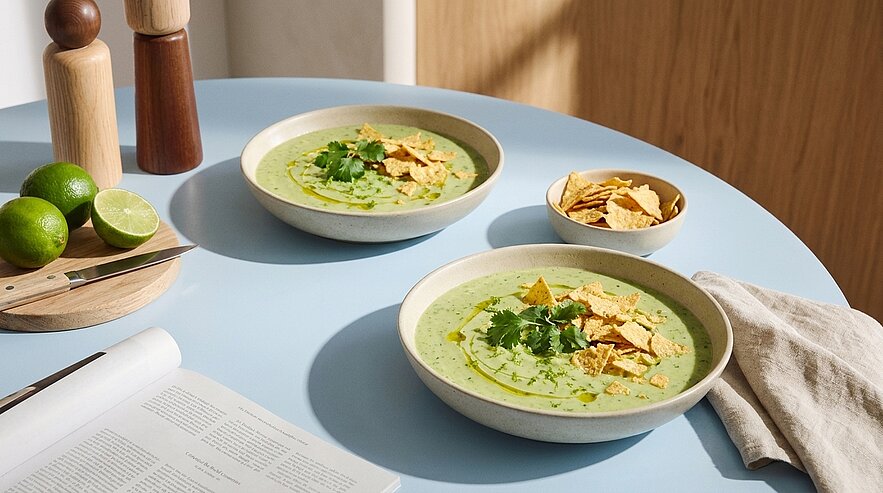 Zwei Schalen cremiger grüner Suppe, garniert mit frischem Koriander und zerbrochenen Tortilla‑Chips, auf einem hellblauen Tisch.