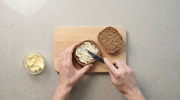 Zwei Hände bestreichen eine Scheibe Vollkornbrot mit Butter auf einem Holzbrett; eine zweite Scheibe liegt daneben, ein kleines Glas Butter steht links.