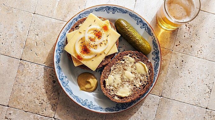 Teller mit zwei Brotscheiben: eine mit Butter, eine belegt mit Käsescheiben und Zwiebelringen; daneben Gewürzgurke, Senfklecks und ein Glas Bier.