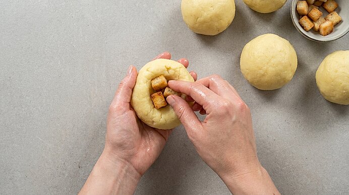 Hände formen einen Teigball und füllen ihn mit kleinen Würfeln (z. B. Brot oder Füllung); mehrere Teigkugeln und eine Schüssel mit Würfeln liegen daneben.