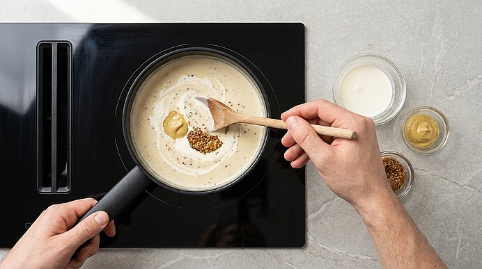 Hände rühren eine cremige Sauce in einem Topf auf dem Herd; mit Holzlöffel werden Senf und Senfkörner eingerührt, Schälchen mit Sahne und Senf stehen daneben.