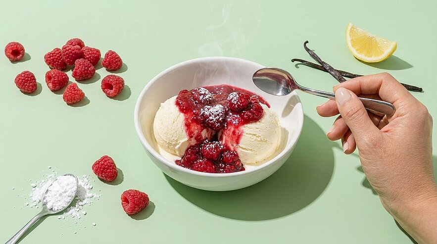 Schale mit zwei Kugeln Vanilleeis, darüber warmes Himbeerkompott; rechts Hand mit Löffel bereit, daneben frische Himbeeren.