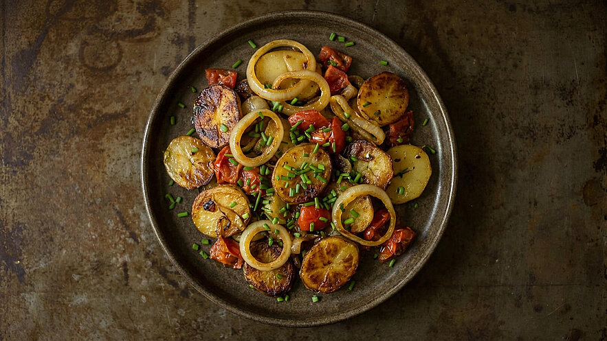 Ein brauner Teller mit gebratenen Kartoffeln, Zwiebelringen und Kirschtomaten. Die Speise ist mit frischem Schnittlauch garniert. Die Kartoffeln haben eine goldbraune Farbe und sind leicht knusprig.