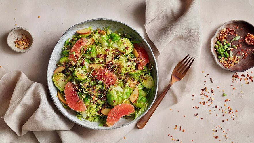 Eine Schüssel mit einem bunten Salat aus Rosenkohl, Apfelscheiben und Grapefruit. Der Salat ist mit Kräutern und Gewürzen garniert. Daneben liegt eine kleine Schale mit Gewürzen und eine Gabel. Der Hintergrund ist neutral und hell.