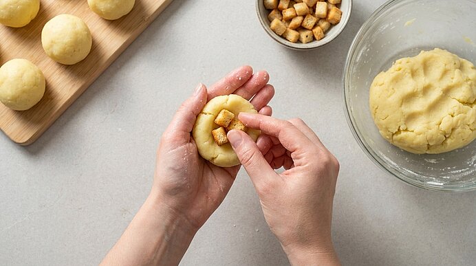 Hände formen und füllen Teigbällchen: ein flacher Teigling wird mit kleinen gewürfelten Brotstücken gefüllt. Daneben eine Schüssel mit Teig und ein Brett mit bereits geformten Teigkugeln.