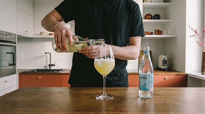 Person gießt ein hellgelbes Getränk aus einer Glasflasche in ein Weinglas auf einer Küchenarbeitsplatte; daneben steht eine Flasche Mineralwasser.