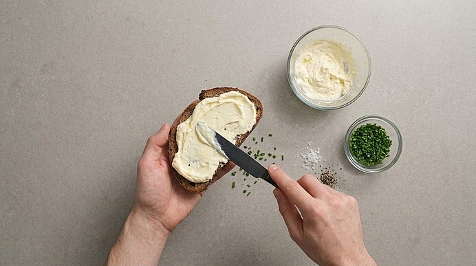 Zwei Hände streichen cremige Sauerrahmbutter auf eine Brotscheibe. Daneben stehen Schüsseln mit Butter und Schnittlauch.