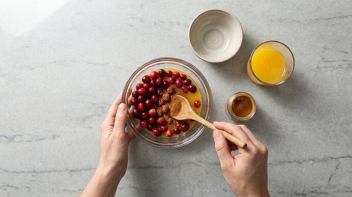 Zwei Hände rühren mit einem Holzlöffel rote Cranberries in einer Glasschüssel; daneben kleine Schalen mit Orangensaft und Gewürz — Zubereitung eines Beerenkompotts.