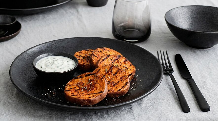 Ein schwarzes Teller mit gegrillten Süßkartoffelscheiben ist im Vordergrund. Neben den Süßkartoffeln befindet sich eine kleine Schüssel mit dipähnlichem, weißen Sauce. Auf dem Tisch liegen ein Messer und eine Gabel. Im Hintergrund sind weitere Geschirrteile sichtbar.