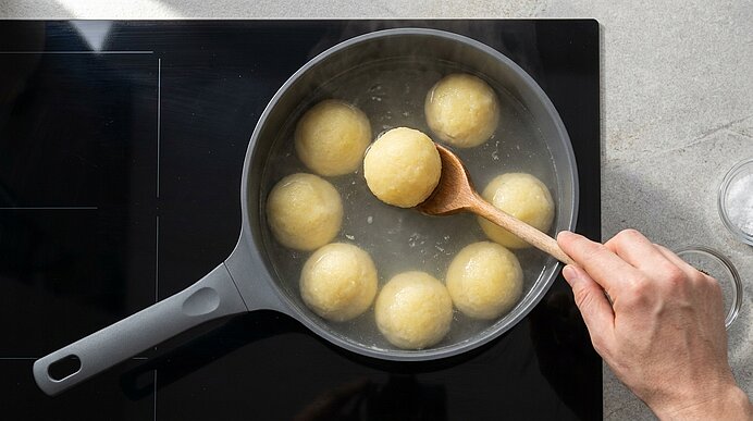 Pfanne auf Herd mit siedendem Wasser und mehreren runden Knödeln; Hand hebt mit Holzlöffel einen Knödel heraus.