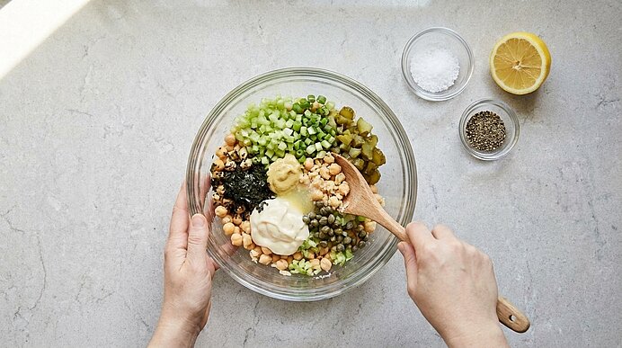 Zwei Hände vermengen Kichererbsen, Gemüse, Mayonnaise und Senf mit einem Holzlöffel in einer Glasschüssel.