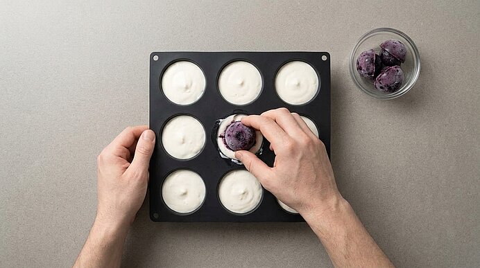 Zwei Hände drücken eine gefrorene Blaubeerkugel in eine mit Vanillemousse gefüllte Silikonform auf grauer Arbeitsplatte.