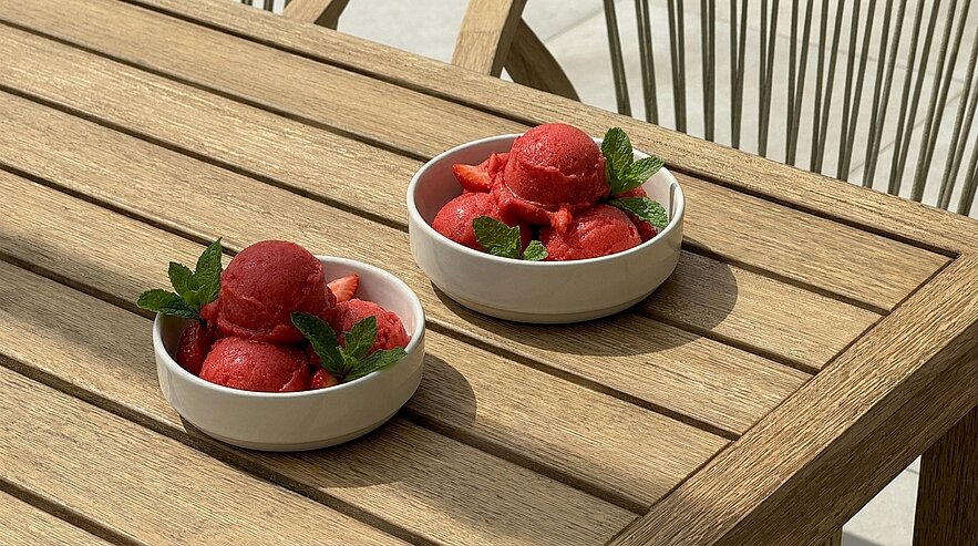 Zwei weiße Schalen mit leuchtend rotem Sorbet, garniert mit frischen Minzblättern, stehen auf einem hellen Holztisch im Freien.