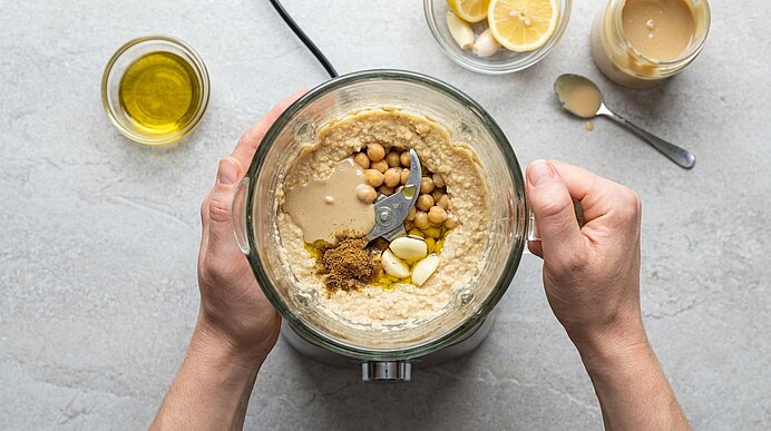 Zwei Hände halten einen Mixerbecher mit Kichererbsen, Tahini, Knoblauch und Kreuzkümmel auf einer grauen Arbeitsfläche.
