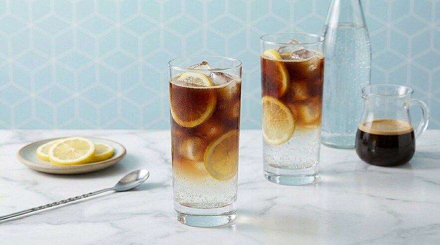 Zwei hohe Gläser mit Espresso‑Tonic: dunkler Espresso über klarem Sprudelwasser, Eiswürfel und Zitronenscheiben als Garnitur.