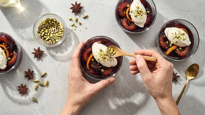 Dessert: Mehrere Glasschälchen mit pochierten Pflaumen in Sirup, jeweils mit einer Kugel Vanillecreme/Eis und gehackten Pistazien; eine Hand löffelt aus einer Schale.