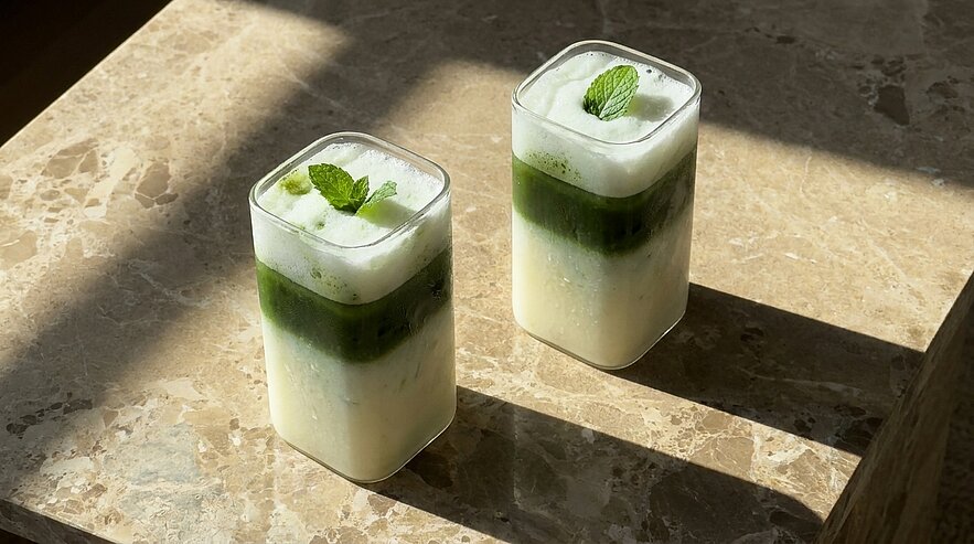 Zwei eckige Gläser mit geschichteten Matcha‑Milchgetränken: milchig weiß, grüne Matcha‑Schicht, Schaumschicht mit Minzblatt, auf Marmor.