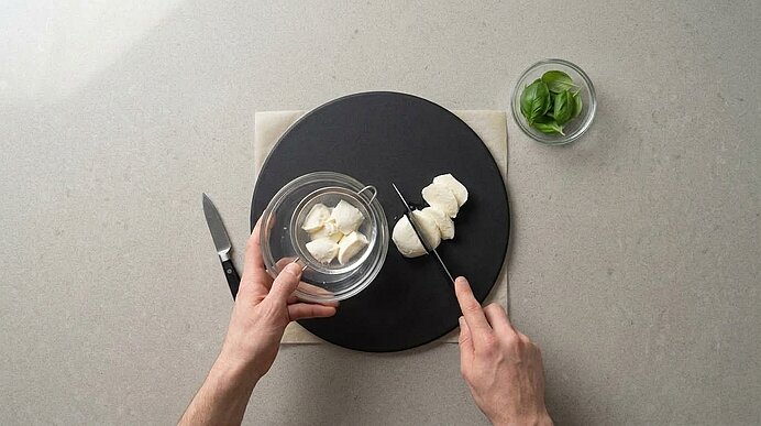 Zwei Hände schneiden Mozzarella auf einem dunklen Brett neben Schüsseln mit weiterem Mozzarella und frischem Basilikum.