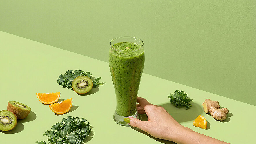 Ein Glas grüner Smoothie wird von einer Hand gehalten. Um das Glas herum liegen halbierte Kiwi, Orangenscheiben, frischer Grünkohl und Ingwerstücke auf einer hellgrünen Fläche.