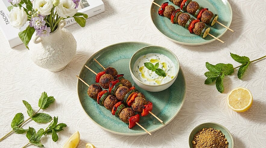 Das Bild zeigt zwei Teller mit Grillspießen, die mit buntem Gemüse und Fleisch belegt sind. Neben den Tellern befindet sich eine Schüssel mit einer Joghurtsauce, garniert mit frischer Minze. Dekorative Elemente wie Kräuter, Zitronenscheiben und eine Vase mit Blumen sind ebenfalls zu sehen.
