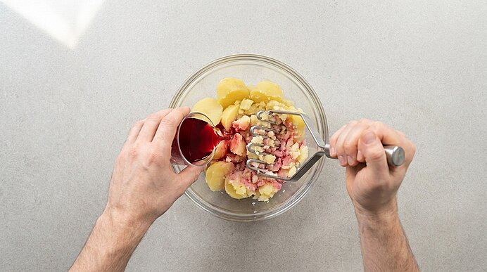 Hände zerdrücken gekochte Kartoffeln in einer Glasschüssel mit Kartoffelstampfer, während aus einem kleinen Messbecher rote Flüssigkeit in die Schüssel gegossen wird.