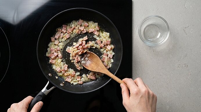 Zwei Hände rühren Speck- und Zwiebelwürfel mit einem Kochlöffel in einer mattschwarzen Pfanne auf dem Kochfeld um.