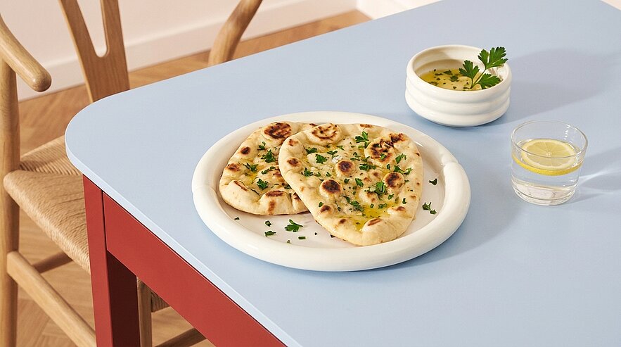 Auf dem Tisch liegt ein Teller mit zwei Stück Fladenbrot, die mit Kräutern garniert sind. Daneben steht eine kleine Schüssel mit einer Sauce und ein Glas Wasser mit einer Zitronenscheibe. Der Tisch hat eine hellblaue Oberfläche.