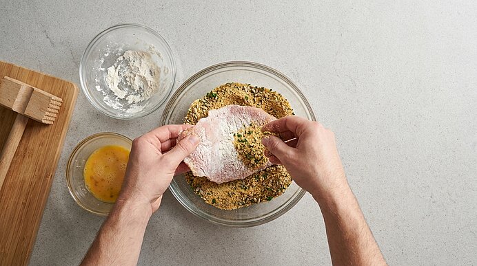 Hände panieren ein Hähnchenschnitzel: zuerst in Mehl, dann in verquirltem Ei und schließlich in einer Kräuter-Semmelbrösel-Mischung in Glasschalen.