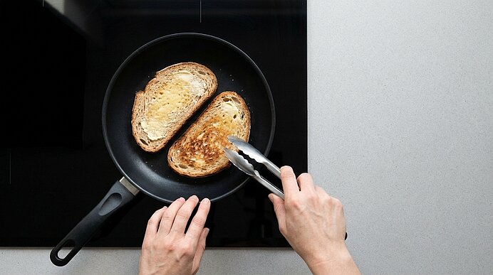 Zwei gebräunte Brotscheiben in einer Pfanne auf dem Herd; eine Hand hält die Pfanne, die andere wendet eine Scheibe mit einer Zange.