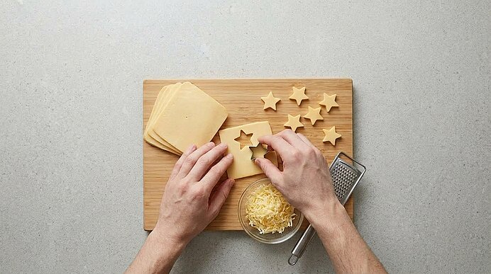 Zwei Hände stechen mit einer Form Sterne aus Gouda auf einem Holzbrett aus. Daneben liegen Käsesterne und geriebener Käse.