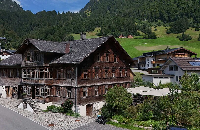 Ein traditionelles Holzhaus mit vielen Fenstern und einem großen, begrünten Garten im Vordergrund. Im Hintergrund sind sanfte Hügel und weitere Gebäude zu sehen. Der Himmel ist klar und sonnig.