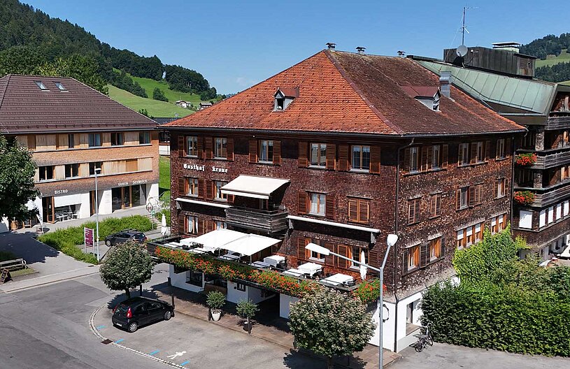 Ein traditionelles Gebäude mit einem rot-bräunlichen Holzfassade, das als Gasthof Krone dient. Vor dem Gebäude sind Tische und Stühle im Freien unter einem Markisenabdeckung. Die Umgebung zeigt grüne Wiesen und Hügel im Hintergrund.