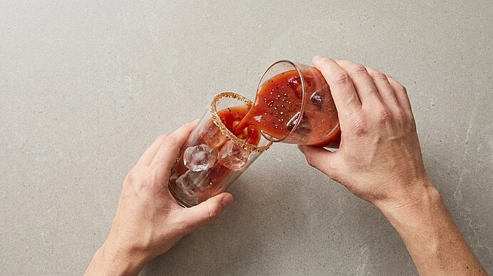 Zwei Hände gießen einen roten Cocktail aus einem Messbecher in ein Glas mit Eiswürfeln und gesalzenem Glasrand.