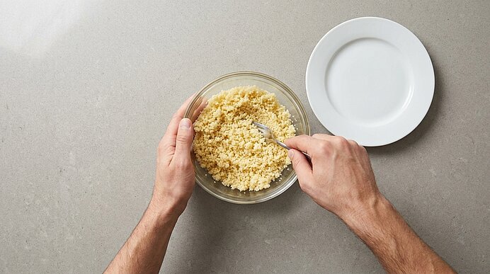 Zwei Hände rühren Couscous in einer Glasschüssel mit einer Gabel; neben der Schüssel steht ein leerer weißer Teller.
