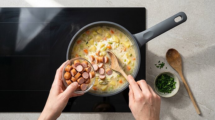 Hände geben geschnittene Würstchen in einen Topf mit cremiger Kartoffel-Gemüse-Suppe auf dem Herd; ein Holzlöffel rührt, gehackte Kräuter in einer kleinen Schale daneben.