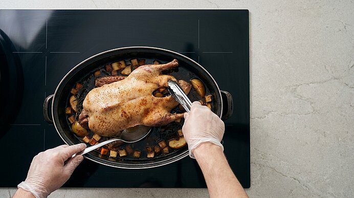 Zwei behandschuhte Hände tranchieren ein goldbraunes Brathähnchen in einem ovalen Bräter, umgeben von gebratenen Kartoffelstücken.