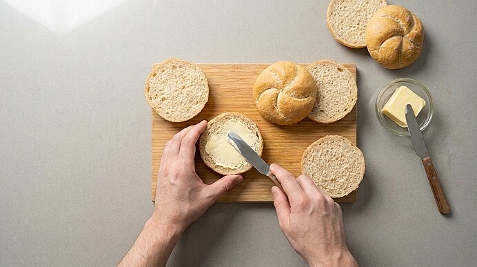 Zwei Hände bestreichen die untere Hälfte einer aufgeschnittenen Semmel auf einem Holzbrett dünn mit Butter.