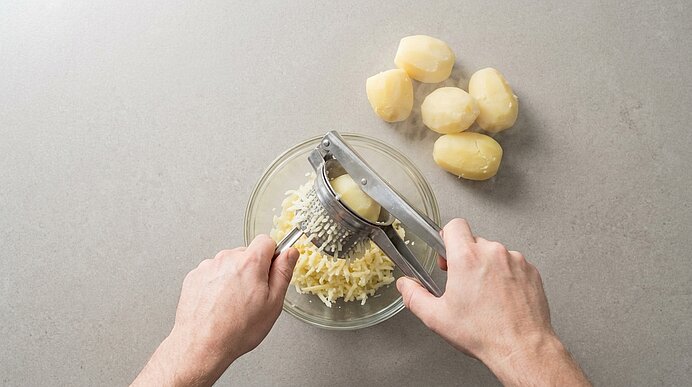 Zwei Hände pressen gekochene Kartoffeln mit einer Kartoffelpresse in eine Glasschüssel; neben der Schüssel liegen vier geschälte, gekochte Kartoffeln.