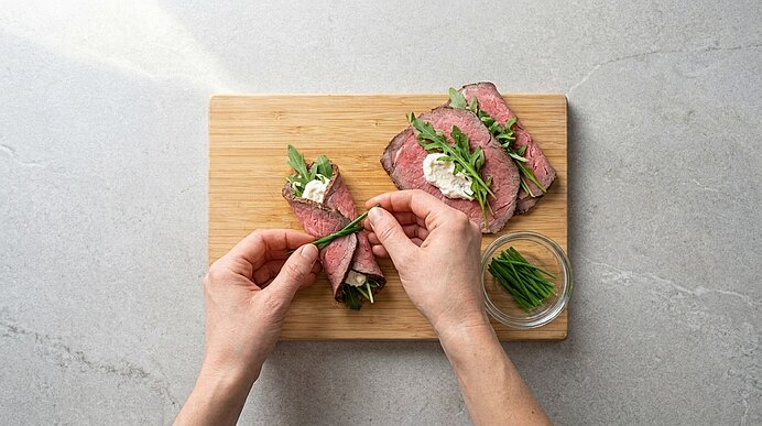 Zwei Hände binden ein aufgerolltes Roastbeef-Röllchen mit einem Schnittlauchhalm auf einem Holzbrett zusammen.